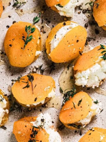 A plate of halved apricots stuffed with white cheese, topped with fresh herbs and cracked black pepper, arranged on a light-colored ceramic dish.