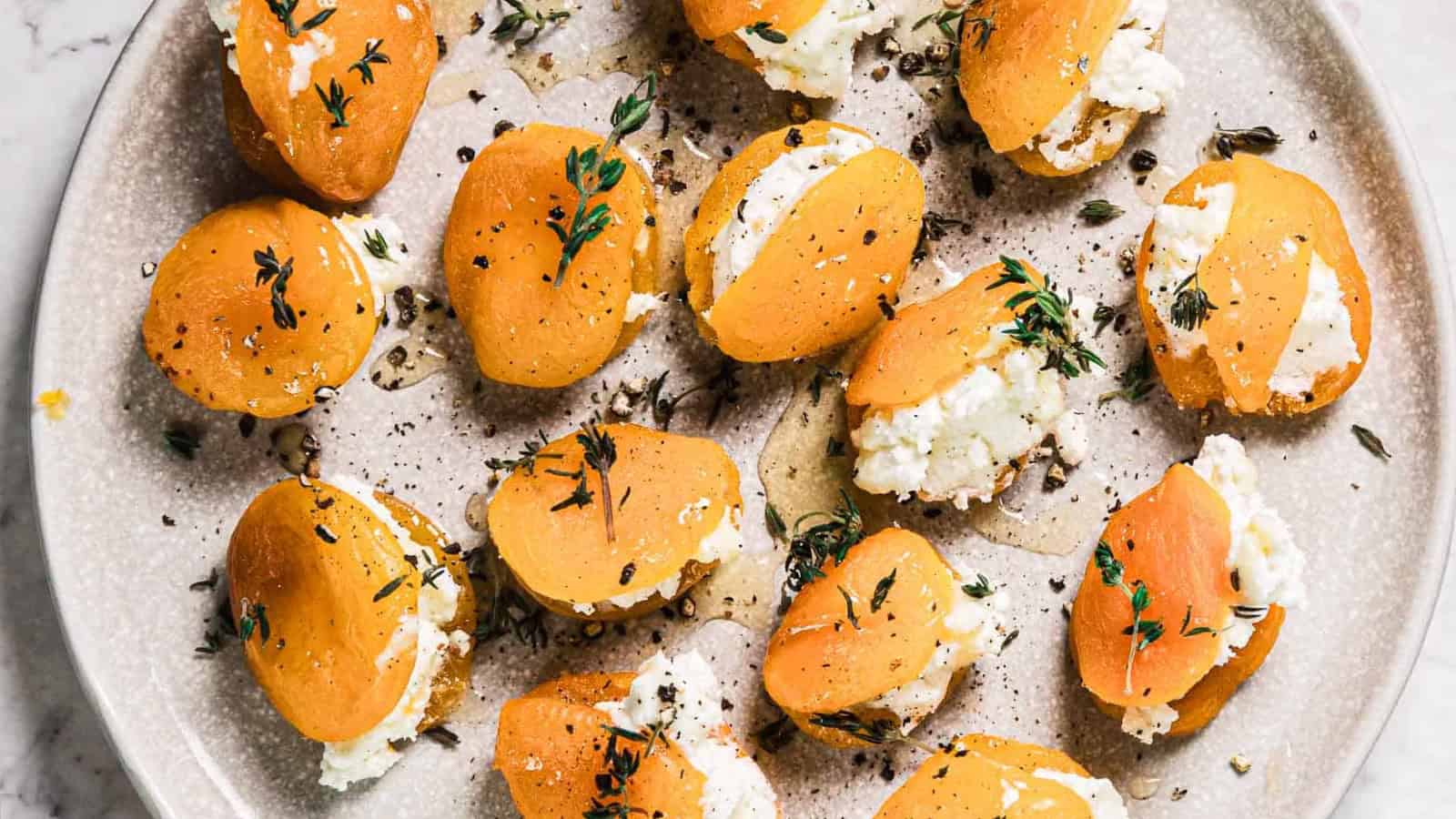 A plate of halved apricots stuffed with white cheese, topped with fresh herbs and cracked black pepper, arranged on a light-colored ceramic dish.