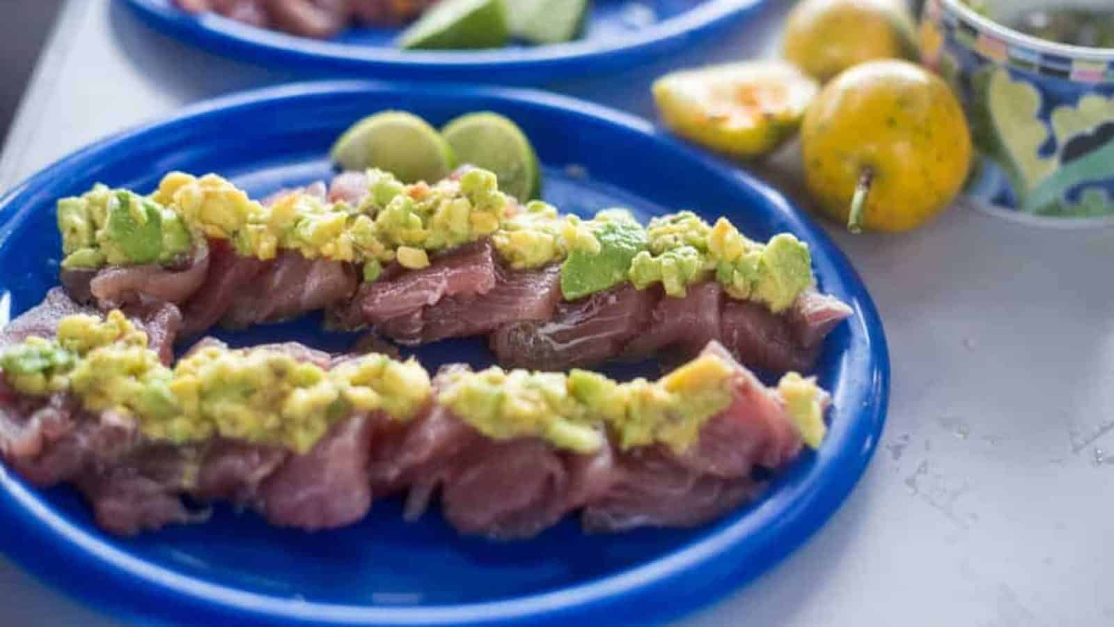 Slices of raw fish topped with diced avocado are arranged on a blue plate, with slices of lime and whole passion fruits visible in the background.