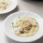 A white bowl filled with creamy soup containing visible chunks of vegetables, topped with ground pepper and a drizzle of oil, set on a light-colored surface. A second bowl is partially visible in the background.