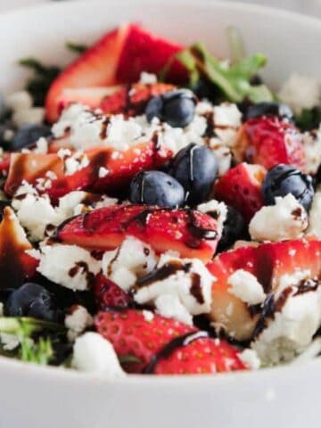 A white bowl filled with salad featuring mixed greens, sliced strawberries, blueberries, crumbled feta cheese, and a drizzle of balsamic glaze, with a spoon resting in the bowl.