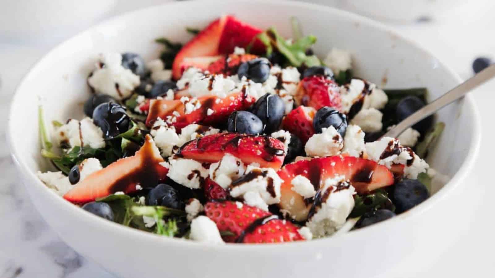 A white bowl filled with a salad of leafy greens, sliced strawberries, blueberries, crumbled white cheese, and a drizzle of dark balsamic glaze, with a metal fork on the right side.