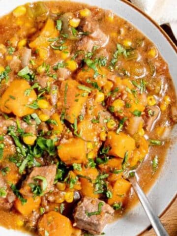 A bowl of stew containing chunks of beef, orange sweet potatoes, corn, and green herbs, served on a white plate with a spoon, sitting on a wooden tray next to a white cloth.
