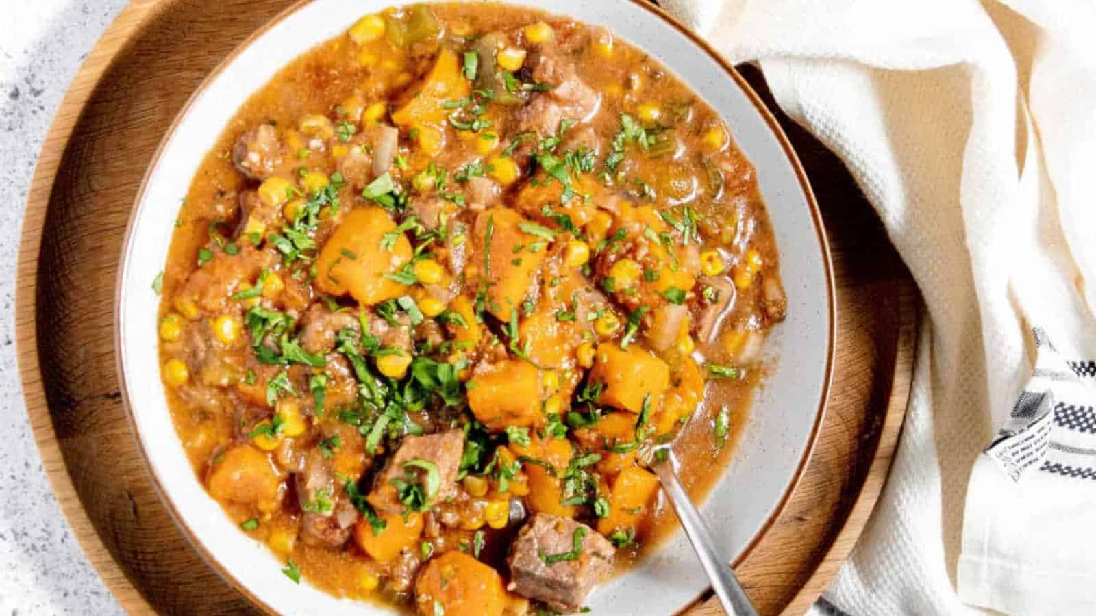 A bowl of beef stew with chunks of beef, carrots, potatoes, and corn, garnished with herbs. A spoon rests in the bowl, and a cloth napkin is nearby on a wooden plate.