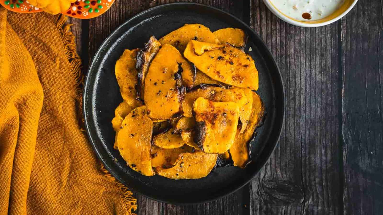 A black plate holds several pieces of roasted or fried yellow pumpkin slices, seasoned with spices.