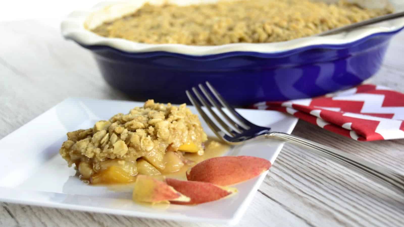 A serving of peach crumble on a square white plate with two peach slices and a fork, placed in front of a blue baking dish containing the remaining crumble. A red and white napkin is nearby.