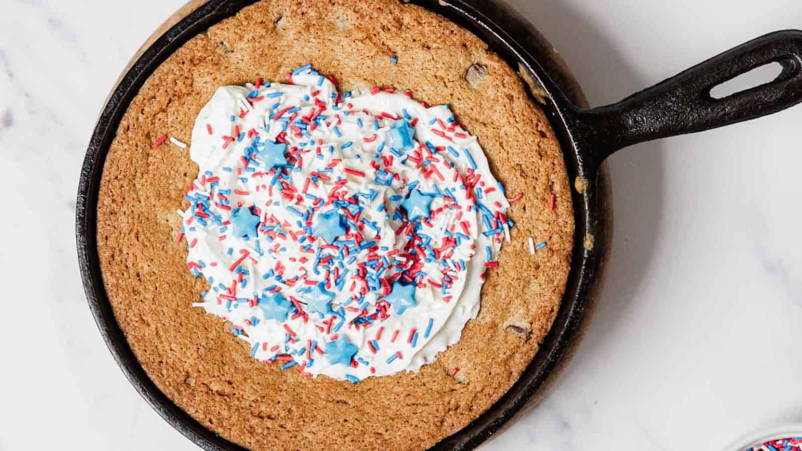 A large chocolate chip cookie baked in a cast iron skillet, topped with white frosting and red, white, and blue sprinkles, viewed from above on a white surface.