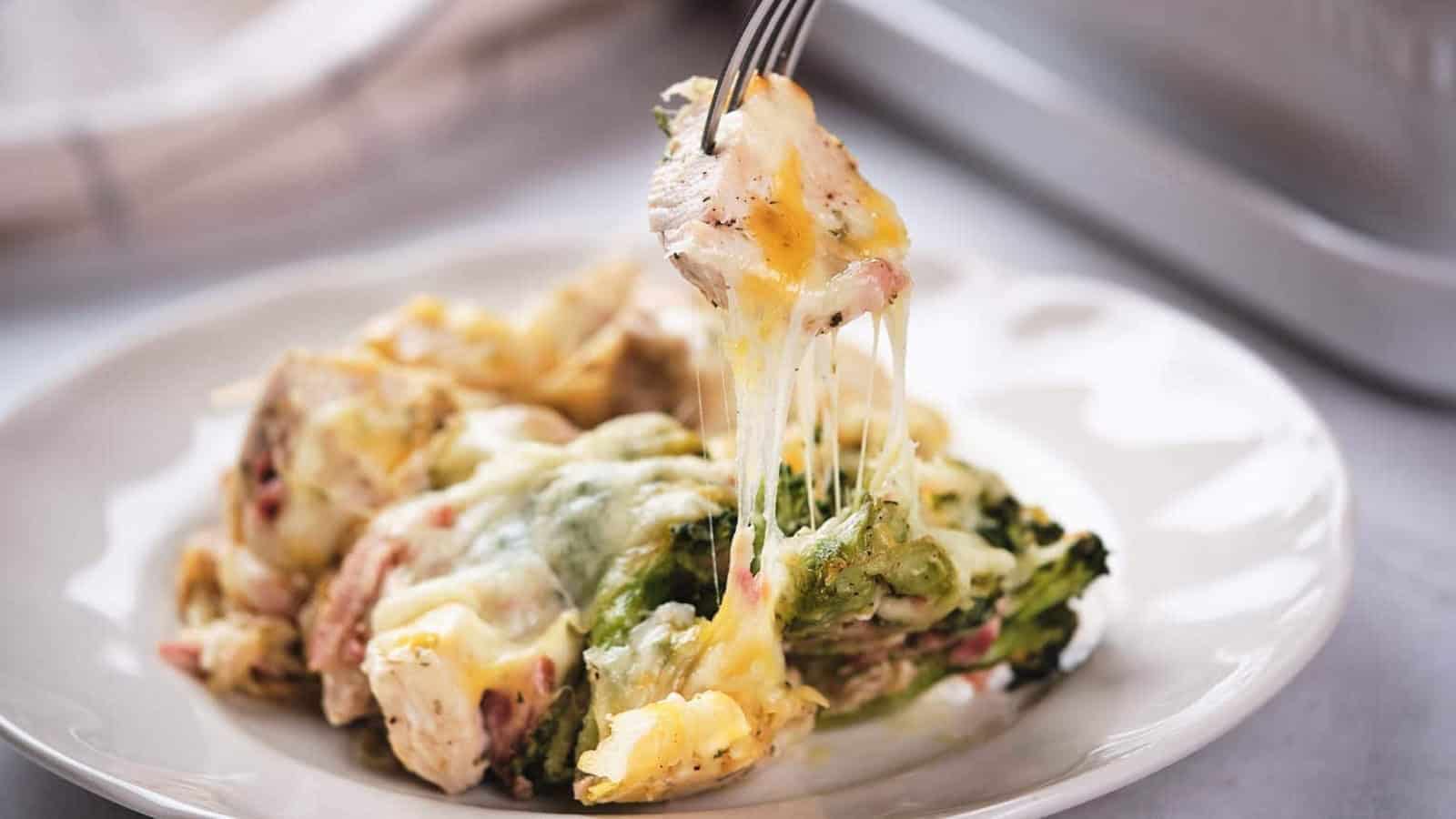 A fork lifts a piece of cheesy baked chicken and broccoli casserole from a white plate. Melted cheese stretches between the fork and the food. The dish contains visible broccoli, chicken, and layers of melted cheese.