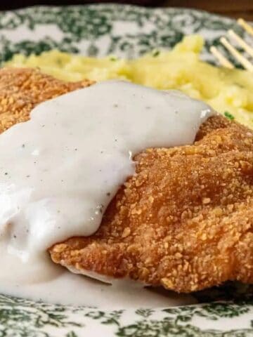A breaded and fried chicken cutlet topped with white gravy is served on a patterned plate alongside mashed potatoes, with a gold fork and knife placed beside the food.
