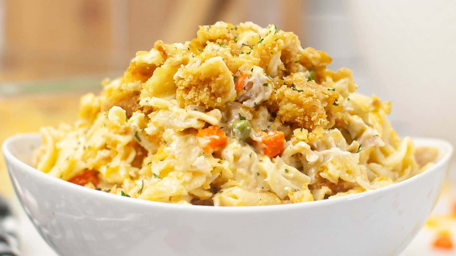 A white bowl filled with creamy pasta mixed with breaded chicken pieces, peas, carrots, and a sprinkling of herbs.