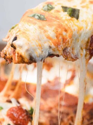 A close-up of a pan of cheesy baked lasagna with a serving being lifted out, showing melted cheese stretching from the pan and bits of fresh basil on top.