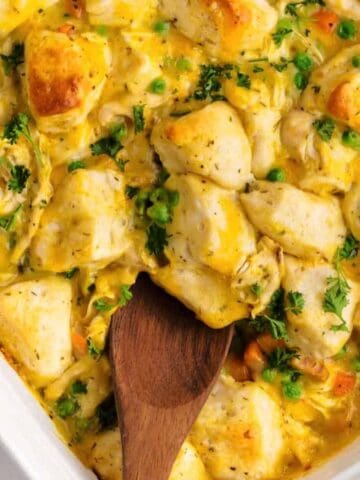A baked casserole dish filled with golden-brown biscuit pieces, chicken, peas, carrots, and a creamy yellow sauce, garnished with fresh parsley. A wooden spoon is resting in the dish.