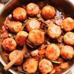 A pan filled with Coq au Vin Chicken Meatballs in a brown sauce with sliced carrots, onions, and herbs. A wooden spoon lifts one meatball from the pan, and the dish is garnished with a sprinkle of chopped herbs.