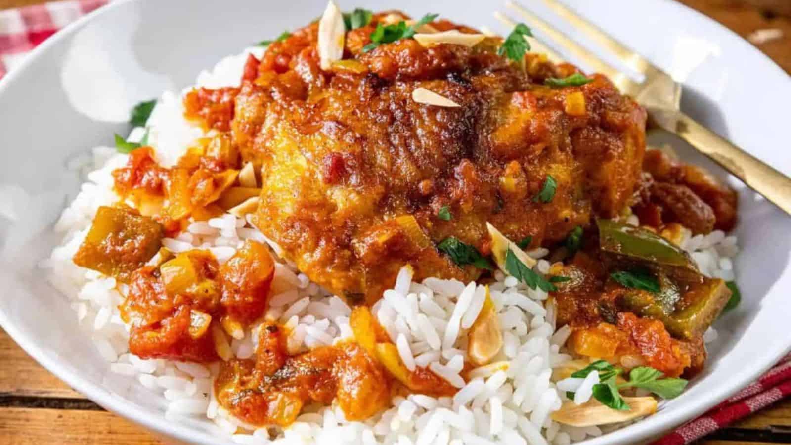 A plate of white rice topped with tender Country Captain chicken in tomato sauce, garnished with chopped parsley and slivered almonds, served with a gold fork on the side.