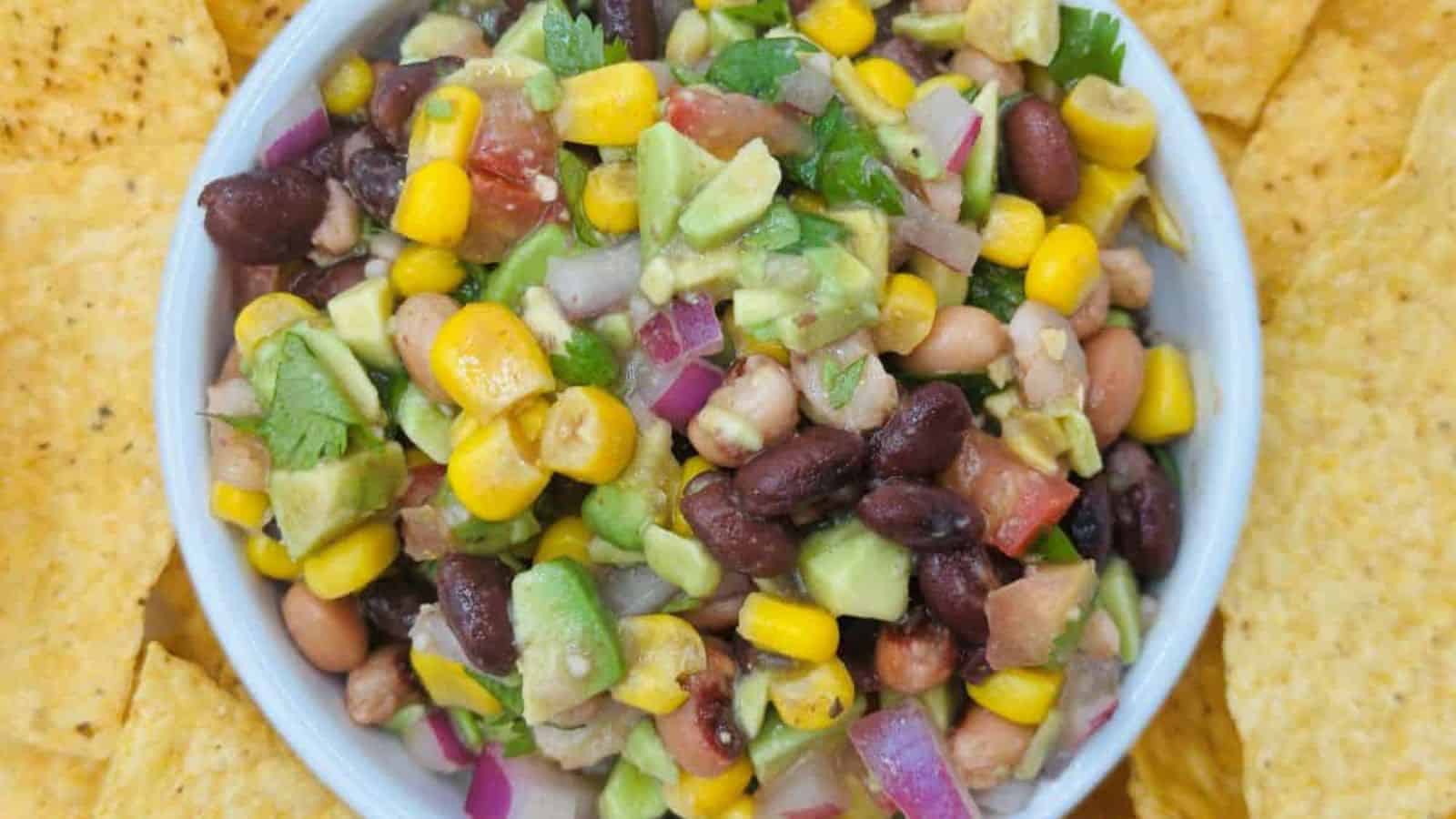 A bowl of bean and corn salad with diced avocado, red onion, tomatoes, and cilantro surrounded by tortilla chips.