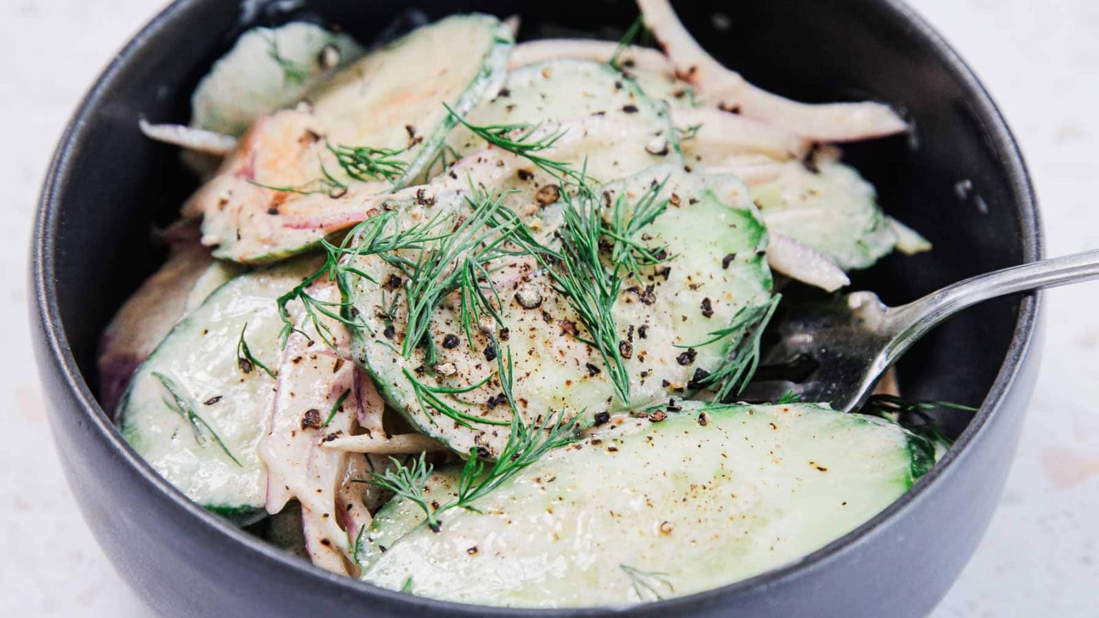 A black bowl filled with creamy cucumber salad, garnished with fresh dill and cracked black pepper, with sliced cucumbers and onions visible. A metal spoon rests inside the bowl.