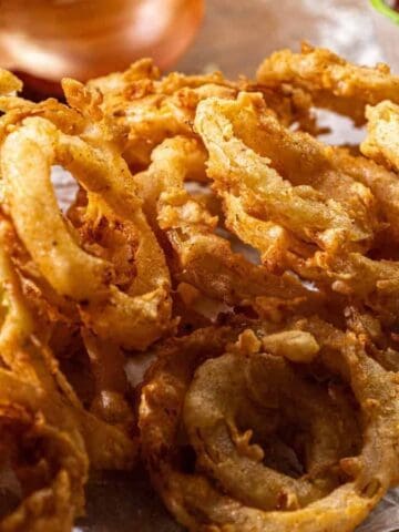 A close-up of crispy, golden-brown fried onion rings on parchment paper with a bowl of ketchup in the background.