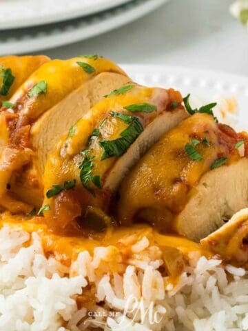 A sliced chicken breast topped with melted cheese, tomato sauce, and herbs is served on a bed of white rice on a white plate. A green and white checkered napkin is in the background.