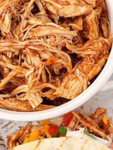 A white bowl filled with shredded, seasoned chicken sits on a patterned plate, next to a taco filled with the same chicken, diced tomatoes, cilantro, and a side dish containing fresh cilantro leaves.