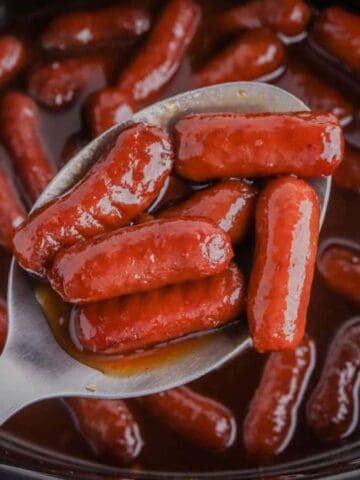 A close-up of a metal spoon holding several glazed cocktail sausages above a slow cooker filled with more sausages in a thick, dark sauce.