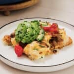A slice of cheesy pizza topped with avocado slices, caramelized onions, chopped chives, and a cherry tomato is served on a white plate with a black rim.