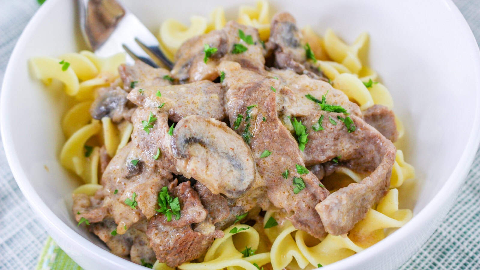 A bowl of beef stroganoff with sliced mushrooms and parsley on top of egg noodles, with a fork on the side.