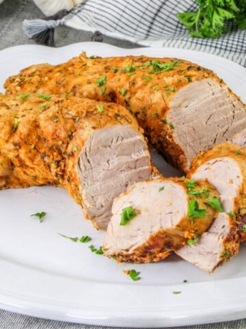 Sliced roasted pork tenderloin garnished with chopped parsley is arranged on a white serving platter, with a sprig of fresh parsley on the side. A gray and white napkin is visible in the background.