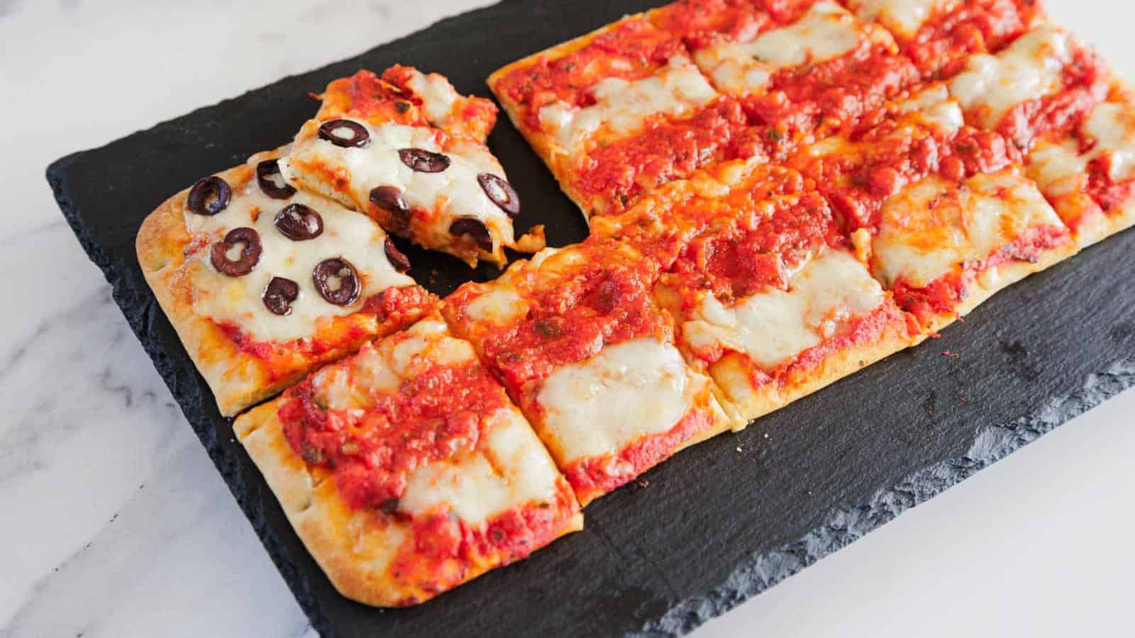 Rectangular pizza cut into square slices on a black slate board, mostly topped with tomato sauce and melted cheese, with one corner slice topped with black olives.
