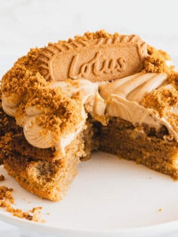 A partially sliced cake with light brown frosting and crumbled Lotus Biscoff cookies on top, displayed on a white cake stand. A blue cup sits in the background.