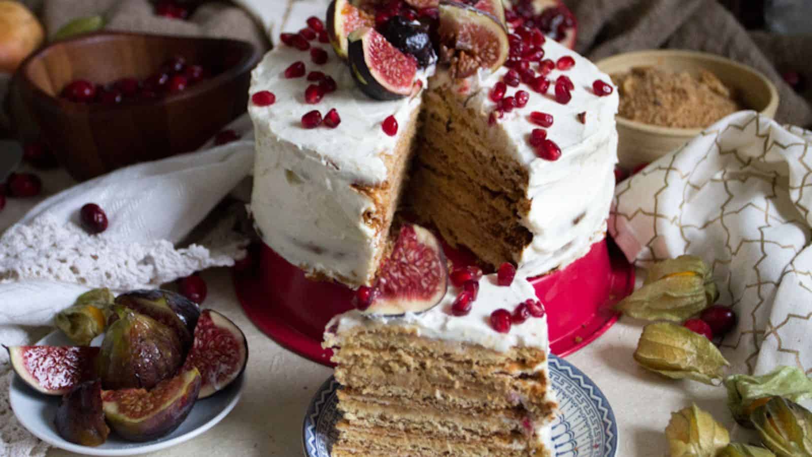 A layered cake with white frosting is topped with figs and pomegranate seeds. A slice is cut and placed on a plate in front. There are bowls of figs, brown sugar, and a white napkin nearby.