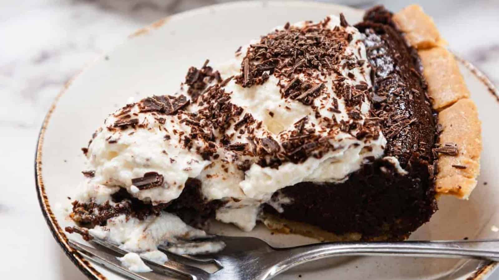 A chocolate cream pie in a metal pie tin, topped with whipped cream and chocolate shavings. A slice has been cut and a metal pie server is visible under the empty space.