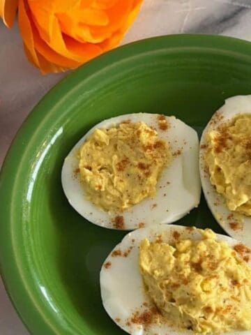 Three deviled eggs topped with paprika are arranged on a green plate. The plate is placed on a light surface next to artificial pink and orange flowers.