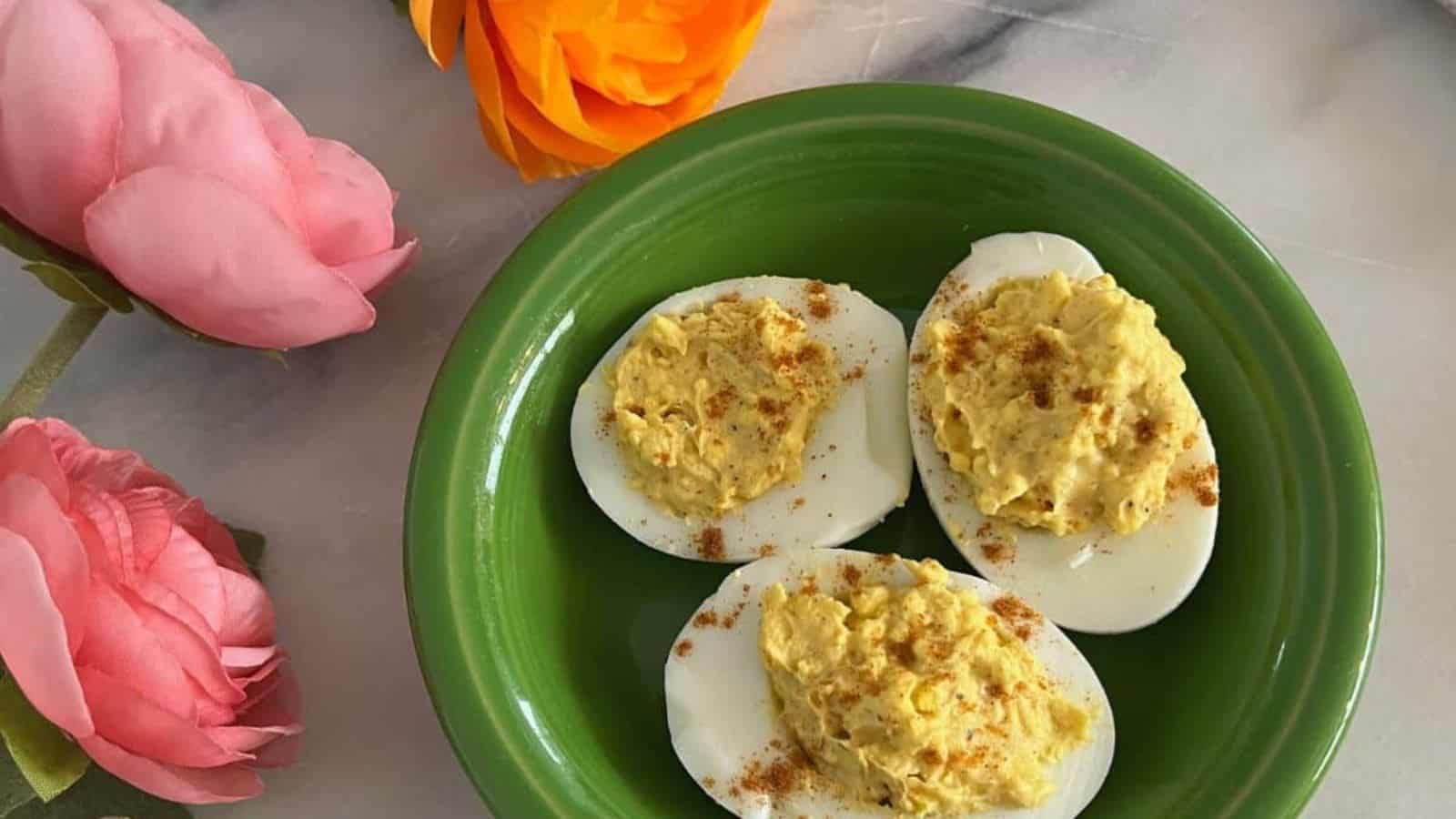 Four deviled egg halves on a green plate, garnished with paprika, accompanied by pink and orange artificial flowers.