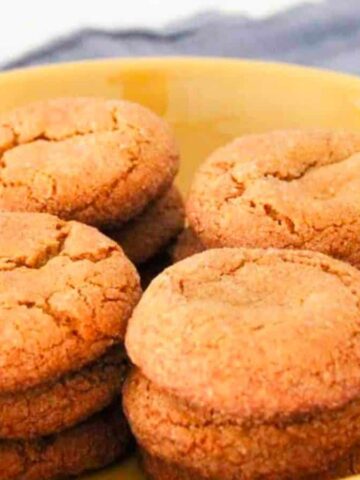 A yellow plate holds two stacks of golden-brown cookies on a blue cloth. The cookies have cracked tops and appear freshly baked. A partially visible jar is in the background.