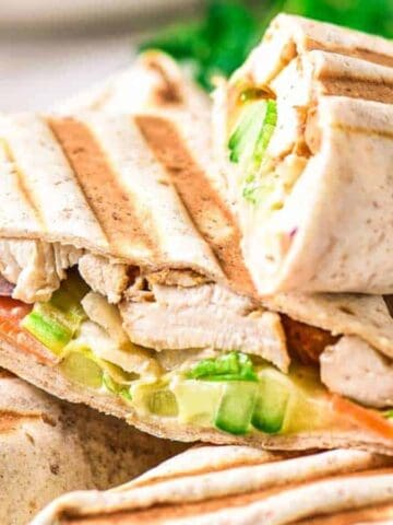 A close-up of two grilled wraps cut in half, showing layers of chicken, cucumber, tomato, lettuce, and other vegetables inside wheat tortillas. Grill marks are visible on the tortilla surface.