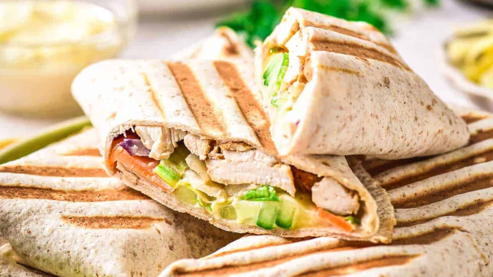 Two grilled wraps are cut in half, showing fillings of grilled chicken, lettuce, cucumber, and other vegetables. The wraps have visible grill marks and are stacked on a plate with a blurred background.