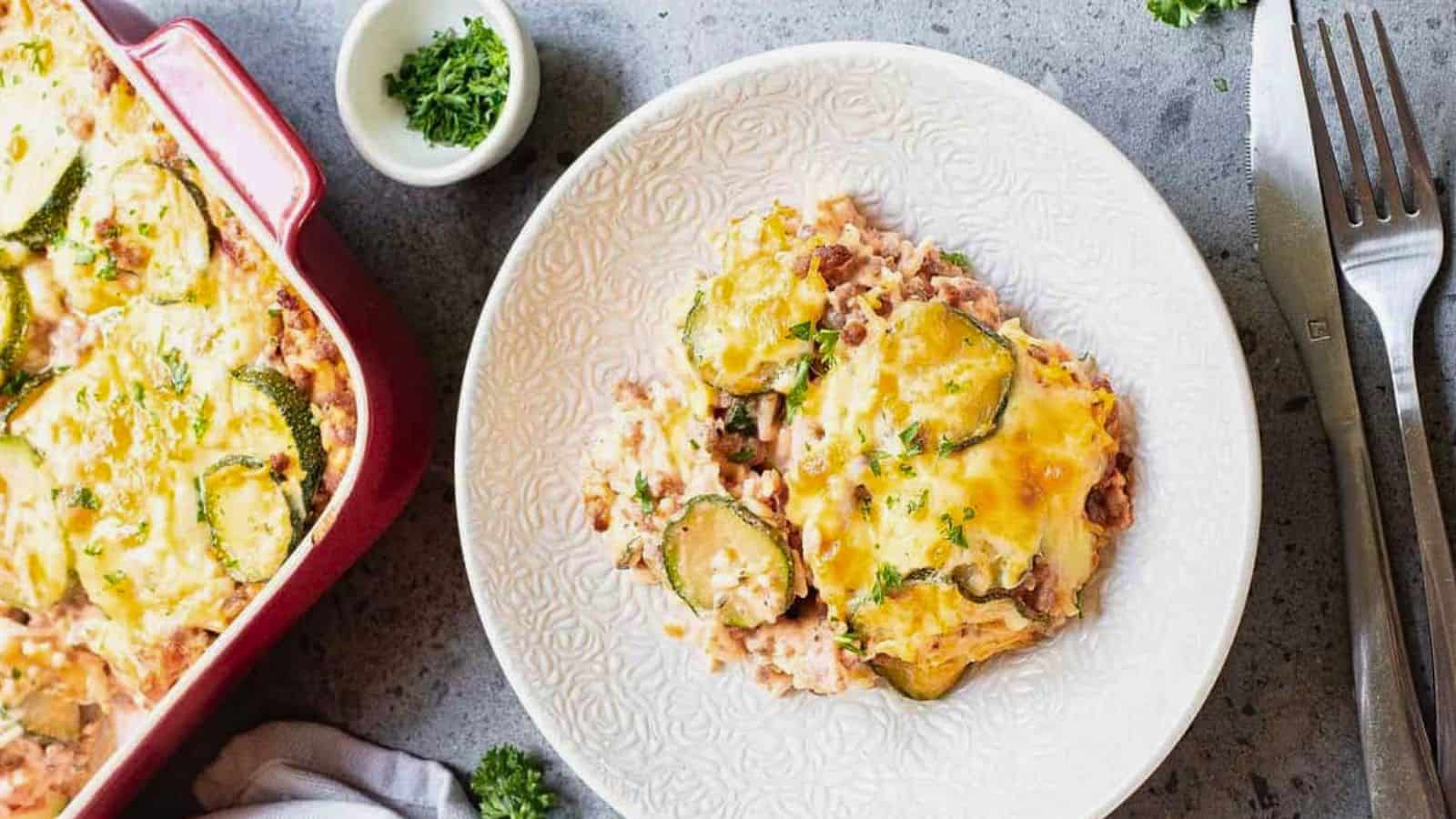 A serving of zucchini casserole with melted cheese is on a white plate next to a fork and knife. The casserole dish is partially visible, and a small bowl of chopped herbs sits nearby&mdash;perfect for fans of classic casseroles.