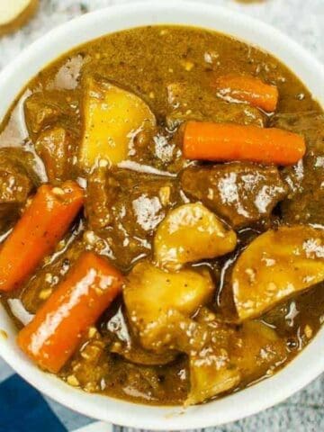 A white bowl filled with beef stew containing chunks of beef, potatoes, and carrots in a brown broth, set on a textured surface with a spoon, napkin, and cut vegetables nearby.