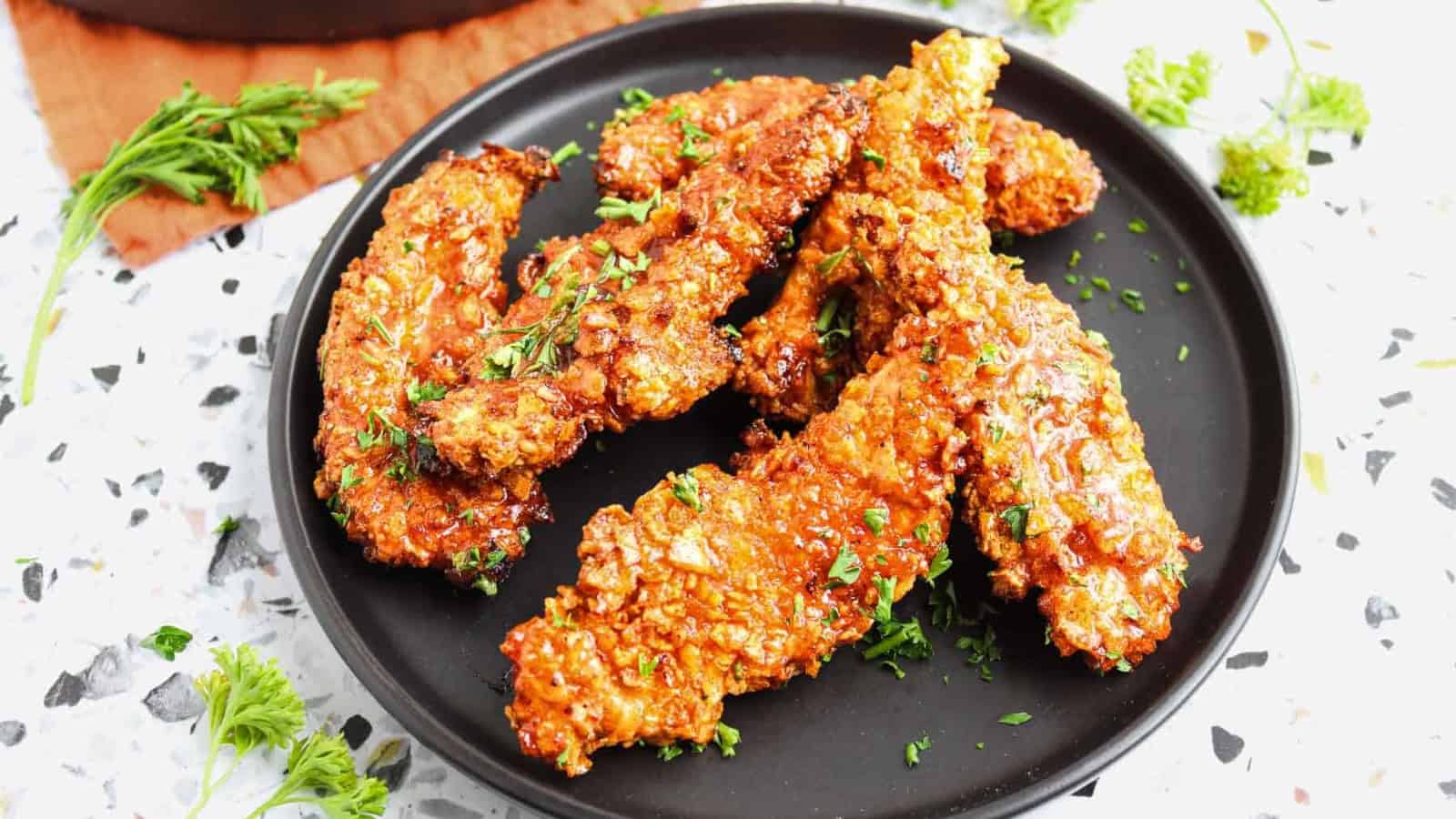 A black plate holding several pieces of golden-brown, crispy fried chicken tenders garnished with chopped parsley, set on a rust-colored cloth with sauce and herbs visible in the background.