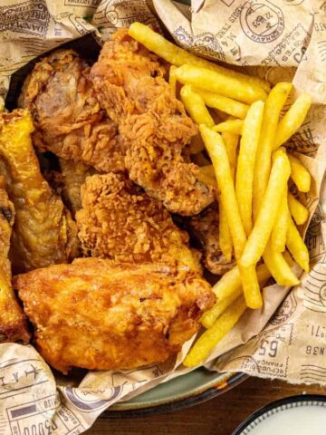 A basket lined with printed paper holds several pieces of fried chicken and a portion of French fries. The basket is on a wooden table alongside plates with seasonings.