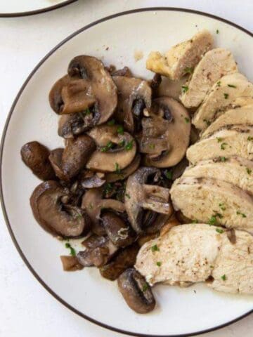 A white plate with sliced cooked chicken breast and sautéed mushrooms, garnished with chopped herbs, sits on a white surface next to a folded white napkin.
