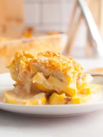 A serving of cheesy yellow squash casserole sits on a white plate, with creamy sauce and a breadcrumb topping. A baking dish with more casserole is visible in the background.