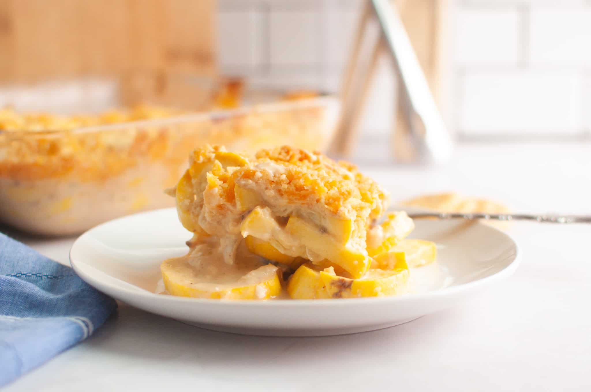A serving of cheesy yellow squash casserole sits on a white plate, with creamy sauce and a breadcrumb topping. A baking dish with more casserole is visible in the background.