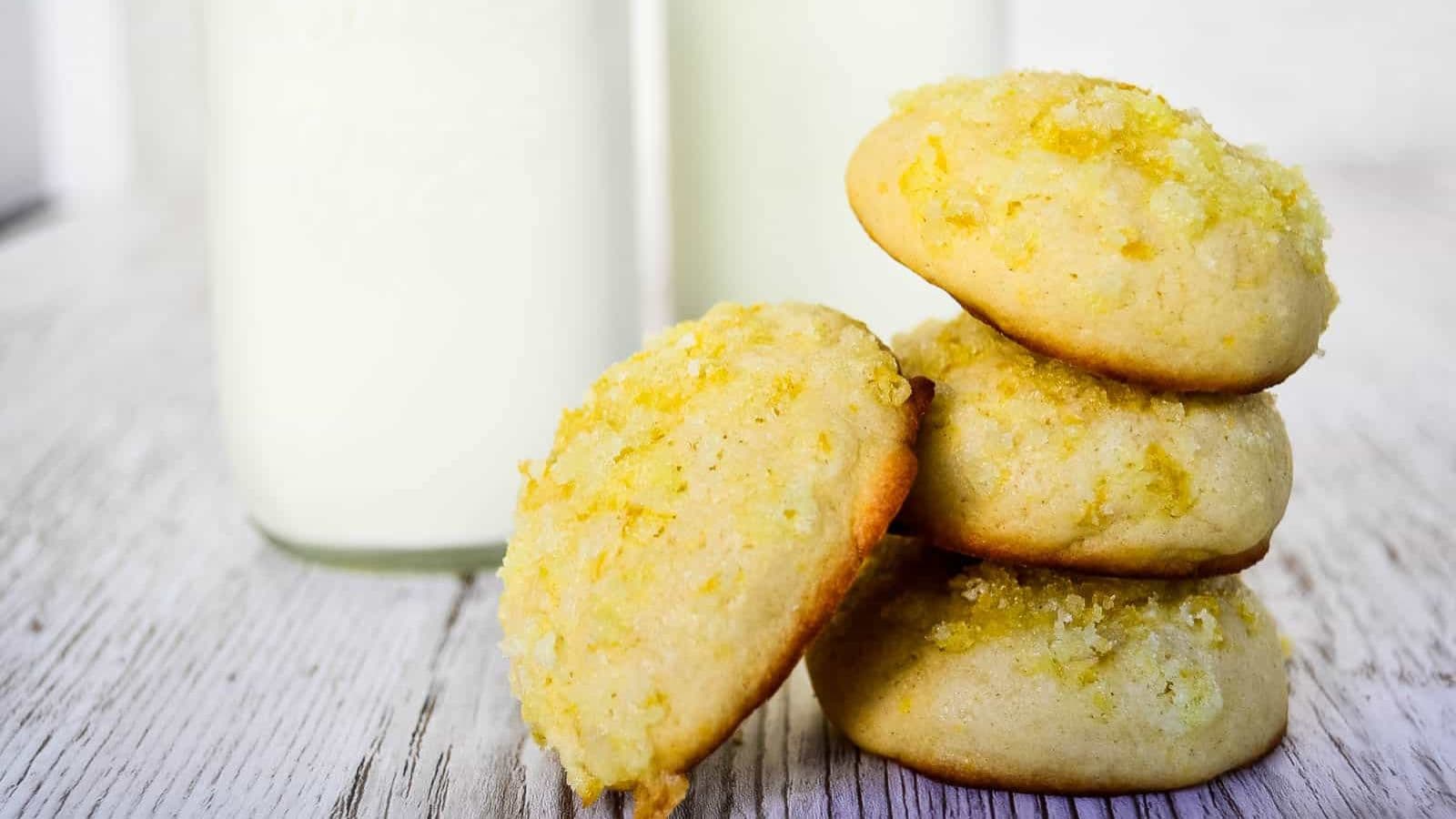 A stack of three lemon sugar cookies with one cookie leaning against the stack, in front of jars of milk.