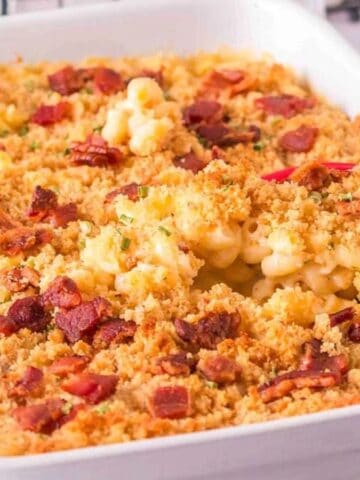 A baked macaroni and cheese casserole topped with golden breadcrumbs and pieces of bacon sits in a white rectangular baking dish. A red serving spoon is scooping out a portion.