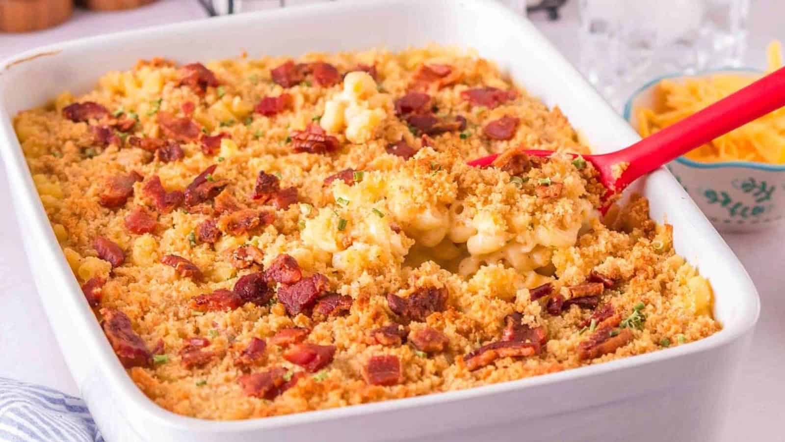 A baking dish filled with macaroni and cheese topped with crispy bread crumbs and bacon bits, with a red spoon scooping out a portion.