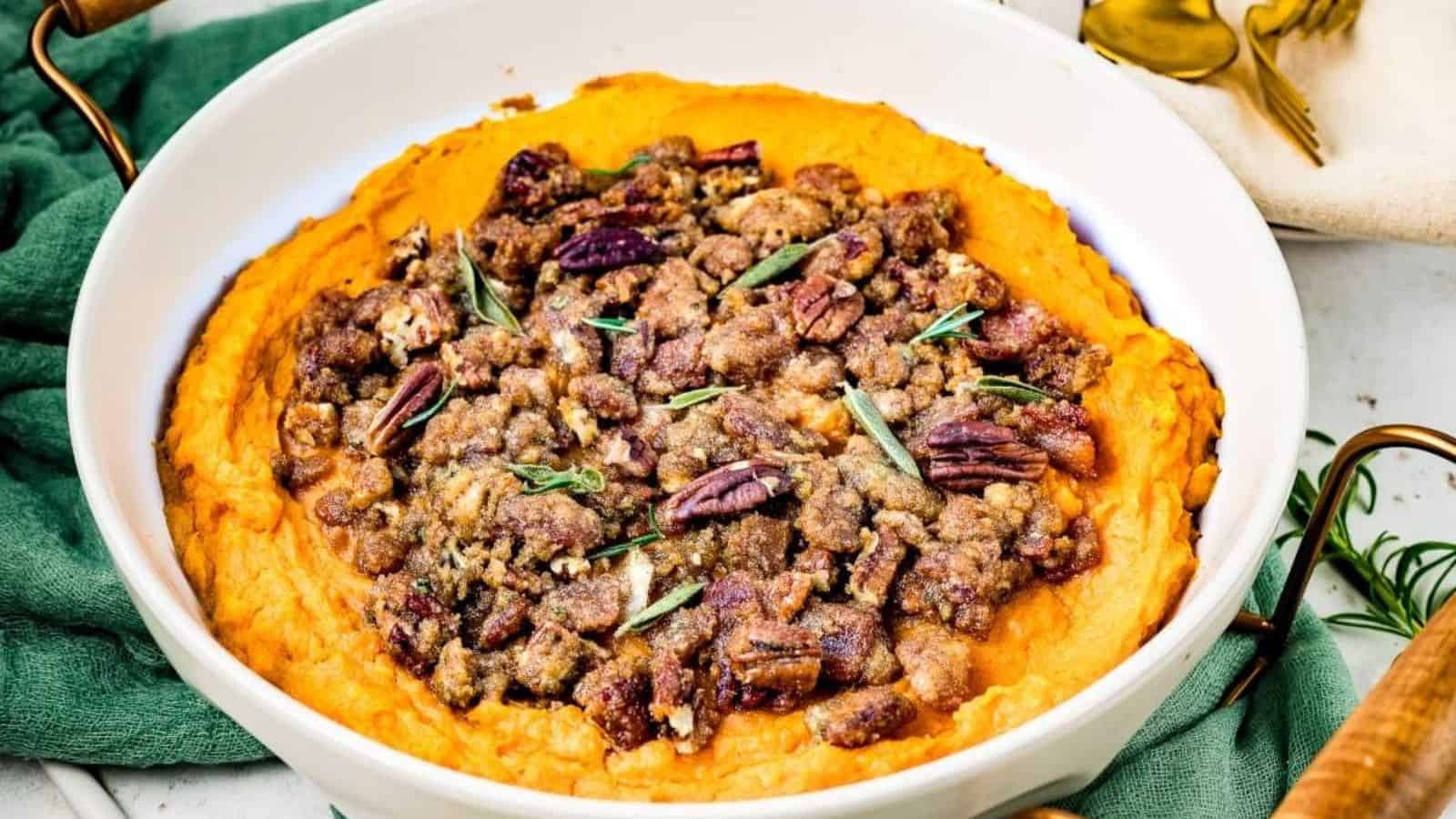 A round dish of sweet potato casserole topped with pecans and herbs, set on a green cloth with serving utensils nearby.