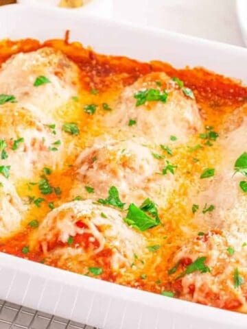 A white baking dish filled with baked stuffed shells topped with melted cheese, tomato sauce, and sprinkled with chopped parsley, sitting on a cooling rack. Plates, forks, and seasoning bowls are in the background.