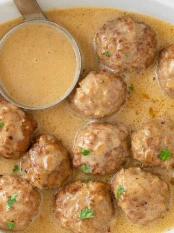 A white oval dish filled with meatballs covered in a creamy brown sauce, garnished with parsley. A metal ladle rests on the edge of the dish, partially submerged in the sauce.