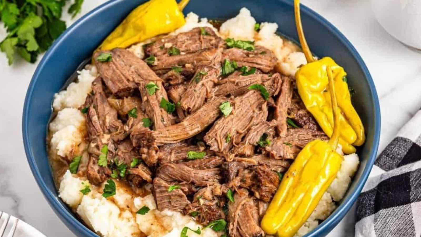 A blue bowl filled with shredded pot roast on mashed potatoes, garnished with parsley and whole yellow peppers.